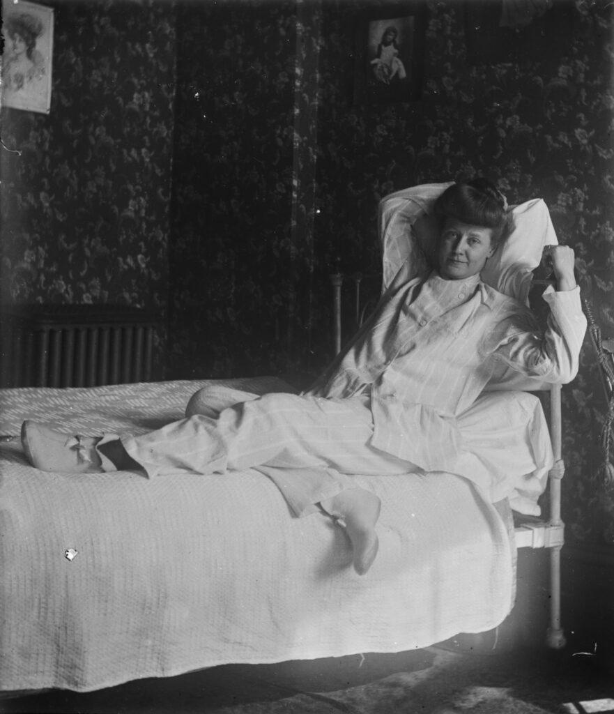 A 1905 photograph depicts a middle-age woman in pajamas reclines in a made bed.